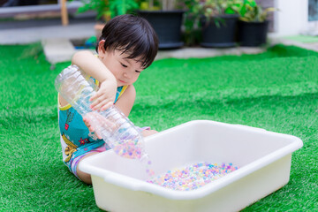 Preschool child boy is using two hands to hold plastic bottle to pour water beads or rainbow beads into white basin. Happy child are sweet smile. Wear blue shirt, yellow border. Toddler 2 years old.