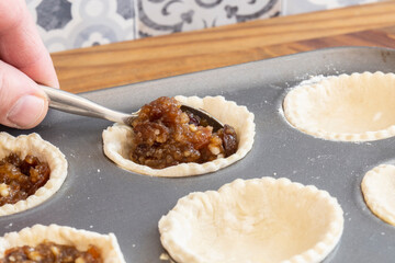 Man filling pastry with mincemeat to make mince pies.  In a metal baking tray on wood