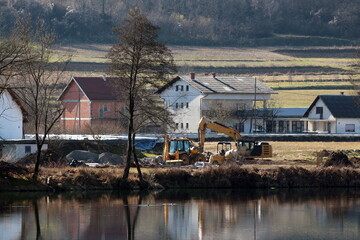 Fototapeta premium Industrial backhoe loader next to compact excavator and small industrial mixer at local levee construction site next to calm river surrounded with construction material and suburban family houses
