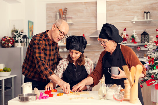 Grandparents helping niece with dessert on christmas day using shape cutter for dogh. Happy cheerful joyfull teenage girl helping senior woman preparing sweet cookies to celebrate winter holidays.