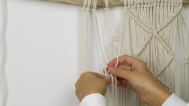 Macrame A woman makes home decor in Boho style. Woman making handmade decor weaves macrame for home decor, white macrame on wood. 4K video close-up of weaving threads into knots.