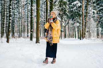 Fototapeta na wymiar Young stylish hipster girl in yellow jacket with a warm scarf walking in the snow forest