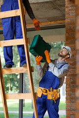 Young workman in overalls and hard hat giving toolbox to his colleague on ladder while working at...