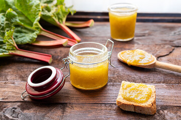 Glass jars of homemade rhubarb jam, a toast with jam and fresh rhubarb stalks on a dark old wooden table near the window, concept of healthy organic breakfast