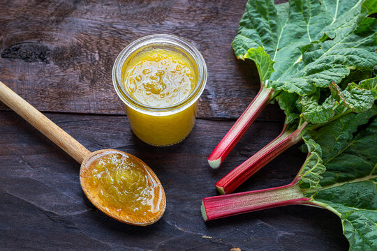 A wooden spoon with homemade rhubarb jam a glass jar and fresh rhubarb stalkson a dark old wooden background, canning food and healthy organic breakfast concept