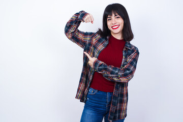 Smiling Young beautiful Caucasian woman wearing plaid shirt against white wall, raises hand to show...