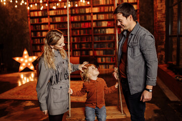 Joyful boy sit on a swing, near stand loving mom with caring dad, wonderful parents have fun with little son, enjoy happy moments together, family winter holidays concept