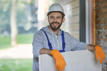 Portrait of handsome young male builder in hard hat looking positive, holding drywall while working...