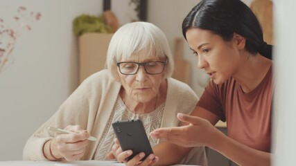 Young Asian female volunteer explaining how to use modern smartphone to senior Caucasian woman and answering her questions while sitting together at home - Powered by Adobe