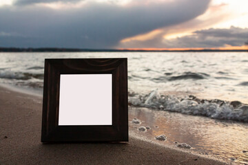 Close-up horizontal photo of a single dark brown wooden squre empty frame for photo on a sea shore with cloudy sky on the background