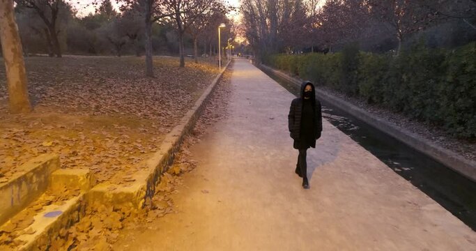 mujer de negro con capucha y mascarilla protecci&oacute;n covit pasea en parque de noche
