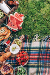Picnic straw basket with healthy food, accessories. Summer picnic with cake, fruits, cheese, wine and snacks on plaid over green background. Top view. Copy space. Summer family lunch. Romantic picnic
