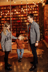 Joyful boy sit on a swing, near stand loving mom with caring dad, wonderful parents have fun with little son, enjoy happy moments together, family winter holidays concept