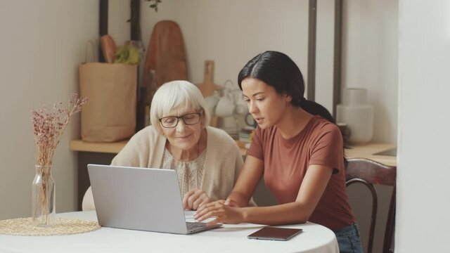 Beautiful Asian female caregiver teaching elderly Caucasian woman how to use the Internet on laptop while sitting together at table in kitchen