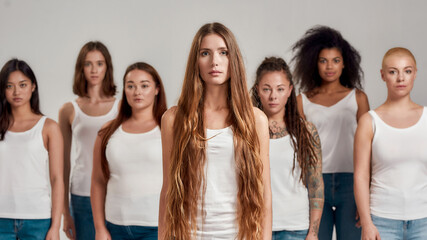 Portrait of beautiful young caucasian woman with long hair in white shirt looking at camera. Group of diverse women posing, standing isolated over grey background