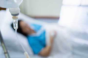 Focus saline IV drip bottle provided to sick woman in bed hospital on the background.