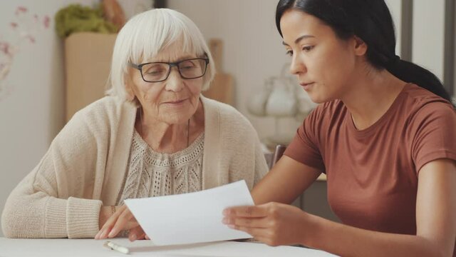 Young female social worker of Asian ethnicity explaining document to elderly Caucasian woman while sitting together at home