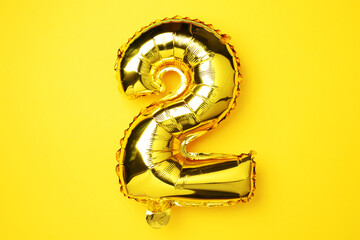 Creative layout. Golden foil balloon number and digit two 2. Birthday greeting card. Anniversary concept. Top view. Copy space. Stylish gold numeral over yellow background. Numerical digit.