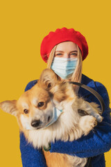 Stylish woman in a mask with a corgi dog in her arms against the background of a yellow wall. Protection against the SARS-CoV-2 virus and prevention in the period of the Covid-19 coronavirus pandemic.