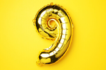 Creative layout. Golden foil balloon number and digit nine 9. Birthday greeting card. Anniversary concept. Top view. Copy space. Stylish gold numeral over yellow background. Numerical digit.