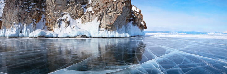 Beautiful landscape of frozen Lake Baikal on a sunny February day. A panoramic view of the icy cliffs of Olkhon Island and clear transparent ice with cracks. Winter holidays, ice travel
