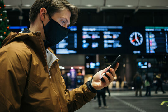Young man in casual outfit stand in middle of train station in face mask on safe social distance from people, use smartphone to check tickets or covid tracing app. New normal concept