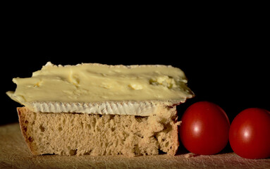 In front of a dark background there is a piece of bread with cheese, a Camenbert, and small tomatoes next to it