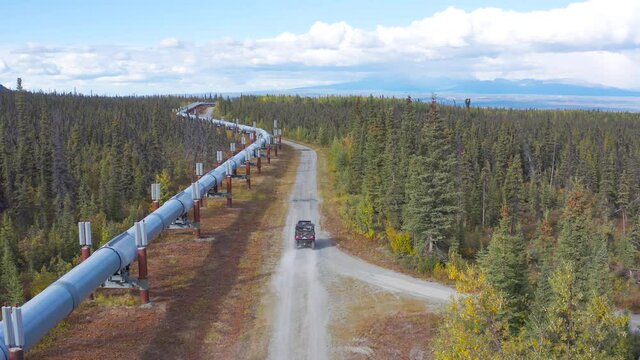 Side-by-side drives along side road of trans-Alaska pipeline in September with fall colors as driver waves hand up to the camera.