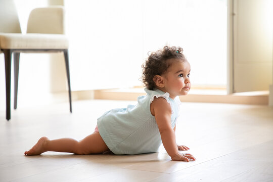 Adorable dark curly haired baby girl wearing pale blue cloth, crawling on floor at home, looking away. Kid at home and childhood concept