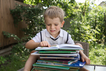 cute toddler boy sits in front of a large stack of children's books, reads intently, on a background of greenery in garden. Focused reading book, development of fantasy, interesting childhood