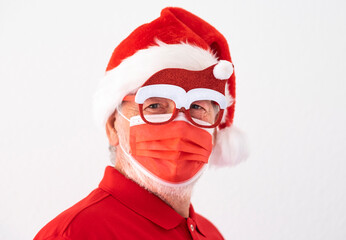 Fototapeta na wymiar Coronavirus and Christmas 2020. Portrait of a senior man with Santa hat wearing surgical mask due to coronavirus and funny glasses. Red color on white background