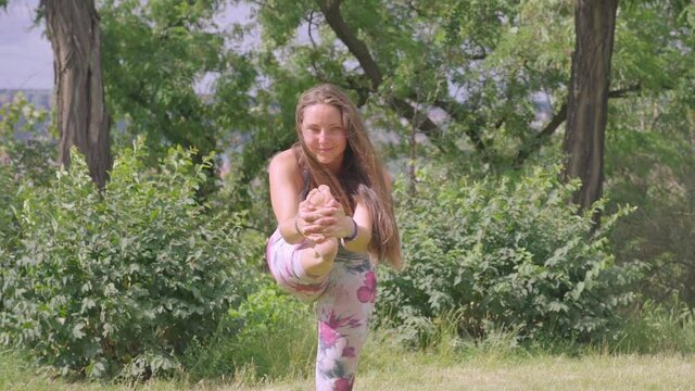Static footage of young woman, performing standing head to knee yoga pose outdoors. Slow motion. Front view.