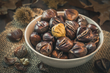 Roasted chestnuts in a bowl, one peeled, on a dark wooden background