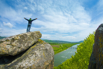 Male hiker standing on top of a cliff. View of the beautiful valley of the mountain river. View from the cliff to the river valley in the mountains. Hikerstanding on rocks with raised hands.