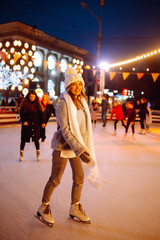 Young  woman ice skating on the ice arena in the city square in winter on Christmas Eve. Winter holiday. New Year magic. Lights around.
