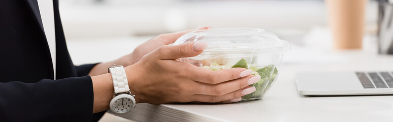 Cropped view of businesswoman holding plastic bowl with food at work on blurred background, 