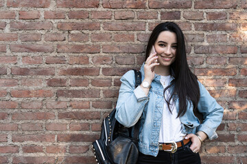 Black-haired female student talking on the phone and leaning against a brick wall