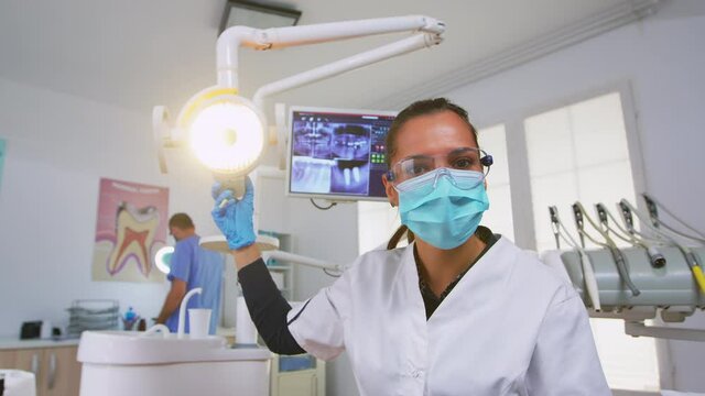 Dentist customizing light over the chair and analysing teeth x-ray before surgery. Dentistry doctor lighting the lamp and examining person wearing protection mask and glasses, nurse helping her