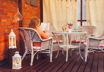 Hygge balcony concept. Woman reading a paper book on home balcony outdoors. Party string lights illuminating the space. Lot of candles and lanterns, curtains hanging with rustic heart party flags.