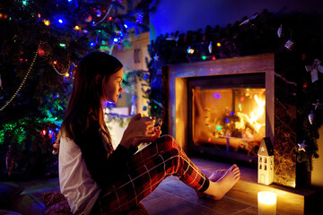 Happy young girl having a cup of hot chocolate by a fireplace in a cozy dark living room on...