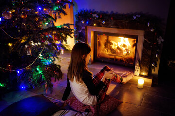 Happy young girl having a cup of hot chocolate by a fireplace in a cozy dark living room on Christmas eve. Celebrating Xmas at home.