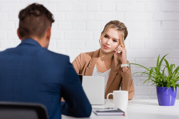  businesswoman looking at colleague, while sitting at table with laptops on blurred foreground