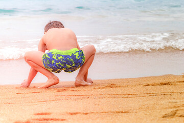 Little brunette boy in blue green shorts doing exercises with squats on the coast of the sea alone....