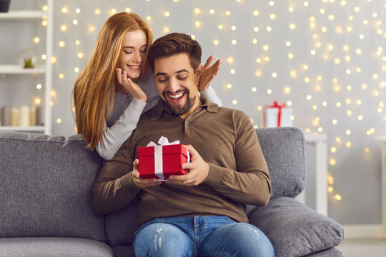 Joyful excited man sits on the sofa in the room and receives a gift from his girlfriend.