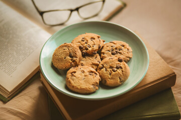 On a stack of old interesting books is a plate of delicious homemade chocolate cookies, and next to it is an open book and glasses, illuminated by light. Hobbies and home comfort.
