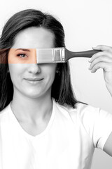 Black and white portrait of young woman painting her eye in yellow color with paintbrush on white...