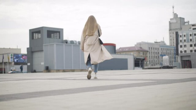 Young Caucasian woman in spring or autumn coat walking away along city square. Back view of confident lady in casual clothes strolling outdoors on cloudy overcast day. Lifestyle concept.