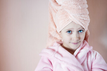 a little girl with blue eyes in a pink robe and a pink towel on her head after a bath smiles and looks into the frame. on a peach background