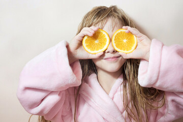 charming blonde girl with wet hair in a pink bath robe covers her eyes with oranges on a pink background
