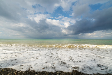 Seascape. Waves roll on the shore against the background of a pre-storm sky with clouds. Water of different colors, white foam.